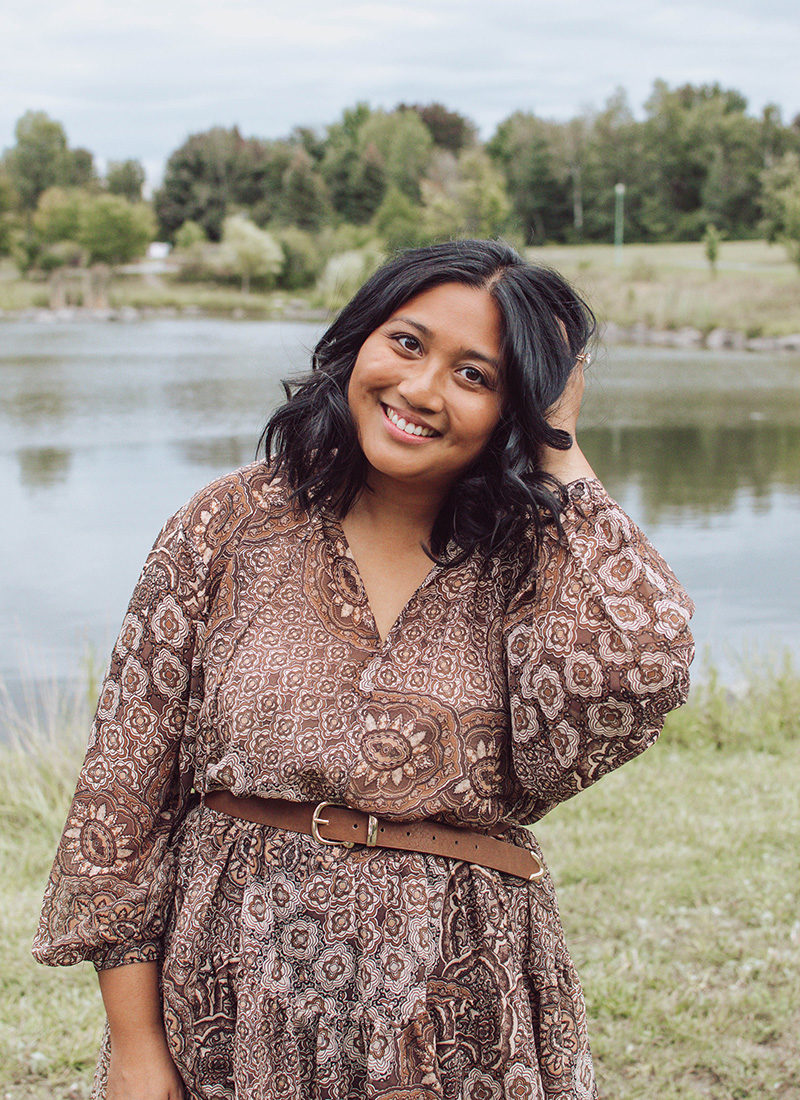 Canadian lifestyle blogger, Lisa Favre, wearing a light, airy brown dress, with curly short hair. Posing in a park in front of a pond.